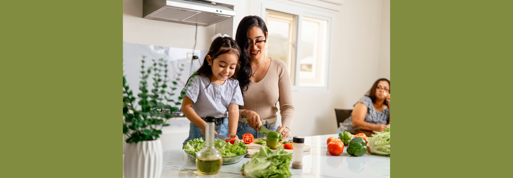 An adult and a young child prepare a fresh salad together in a bright kitchen, chopping vegetables on a countertop filled with lettuce, tomatoes, and peppers, while another adult sits nearby in the background.